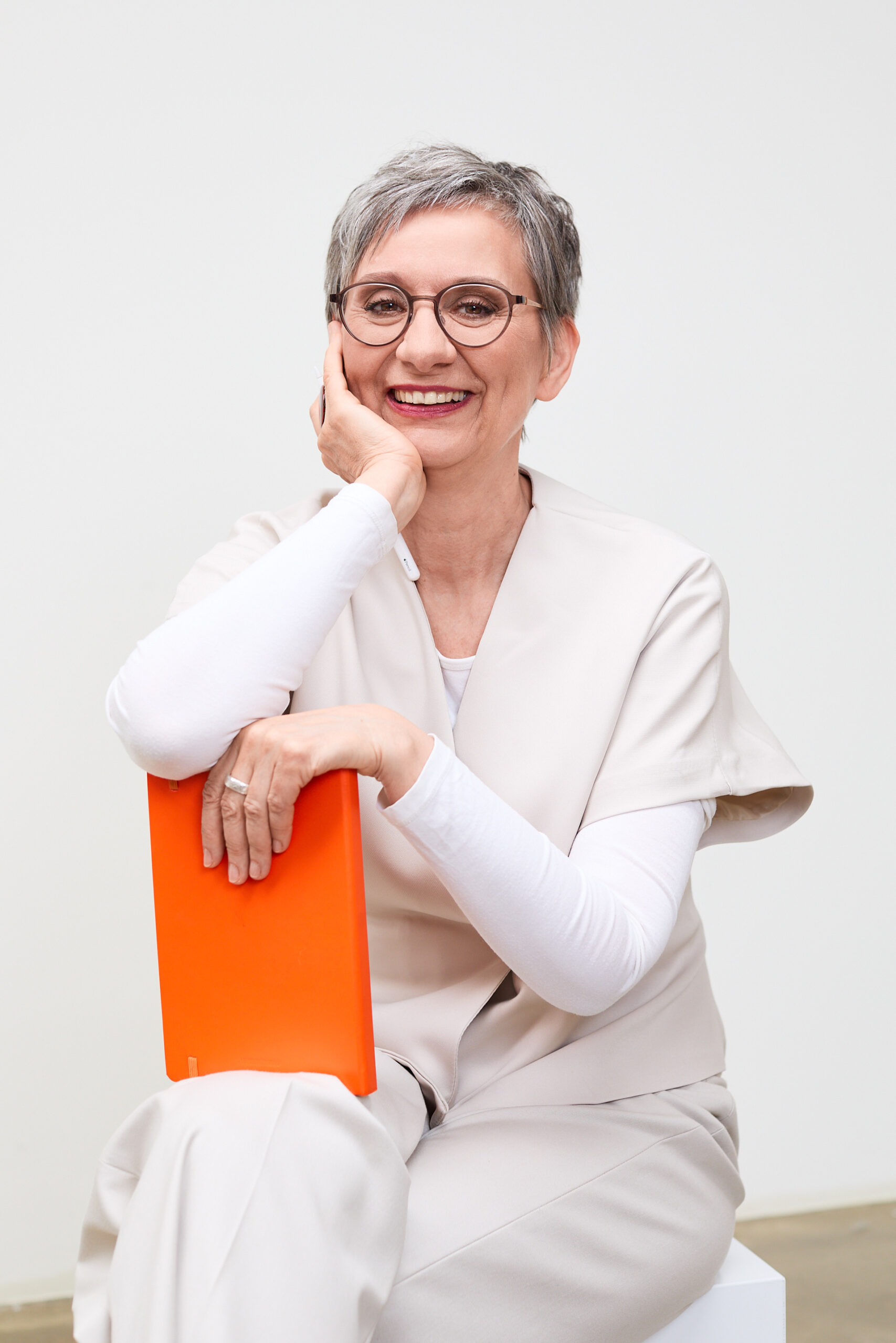 Personal-Branding-Shooting Eva Kräling – sitzend mit Kinn auf der Hand, orangefarbene Mappe im Arm